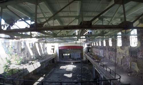 Abandoned industrial space with a large open area, broken windows, and remnants of machinery, illuminated by natural light.