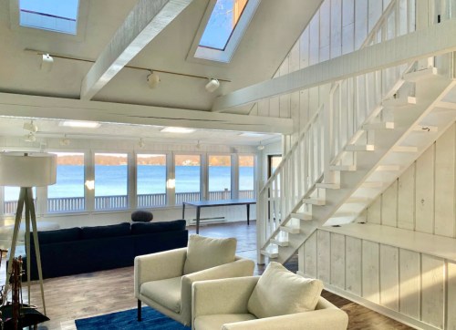 Bright, airy interior with white walls, a staircase, and large windows overlooking a lake. Cozy seating area included.