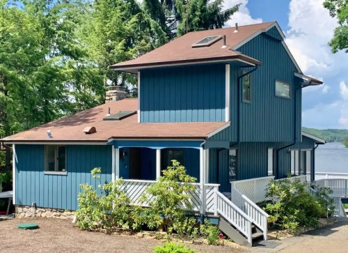 A blue two-story house with a brown roof, surrounded by greenery and a clear blue sky.