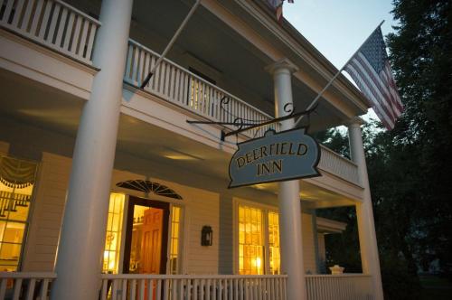 A charming inn with a sign reading "Deerfield Inn," illuminated windows, and American flags on a porch at dusk.