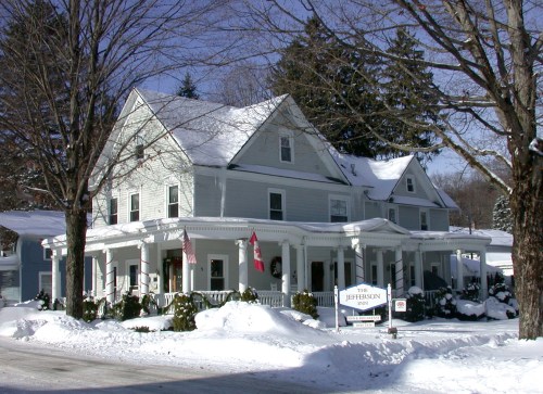 A large, historic house with a snow-covered yard, featuring a sign that reads "The Jefferson" and flags on the porch.
