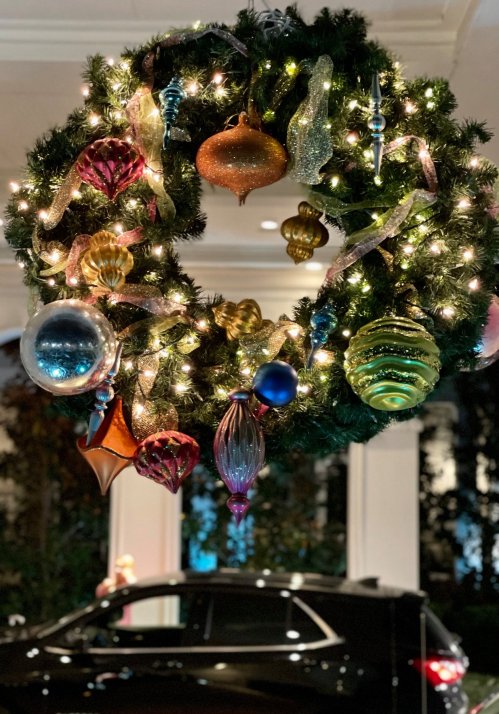 A festive holiday wreath adorned with colorful ornaments and lights, hanging above a parked car.