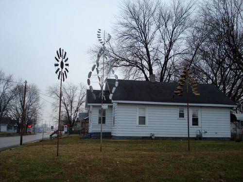 A white house with a gray roof, featuring decorative windmills and bare trees in a cloudy sky.
