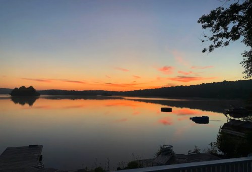 A serene lake at sunset, reflecting colorful skies and silhouettes of trees and boats on the calm water.