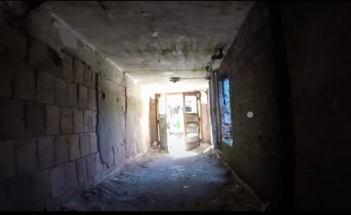 A dimly lit corridor with crumbling walls, leading to a bright doorway at the end. Dust and debris are visible on the floor.