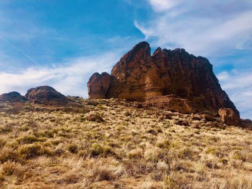 A rugged rock formation rises above a grassy landscape under a blue sky with wispy clouds.