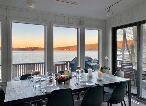 A dining table set for a meal, with large windows overlooking a serene lake at sunset.