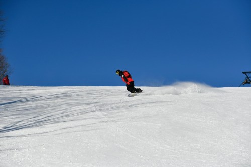 A snowboarder in a red jacket glides down a snowy slope under a clear blue sky.