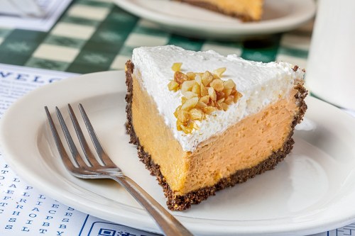 A slice of pumpkin cheesecake topped with whipped cream and nuts on a white plate, with a fork beside it.