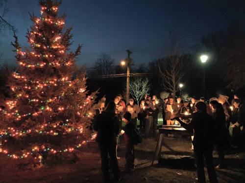 A crowd gathers around a brightly lit Christmas tree at dusk, holding candles in a festive outdoor setting.