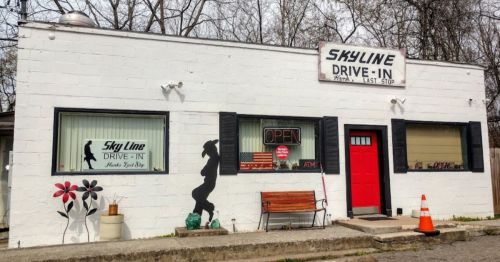 A small, white building with a red door, featuring a drive-in sign, open window, and decorative flowers on the wall.