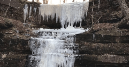A frozen waterfall with icicles hanging from rocky cliffs, surrounded by bare trees and a winter landscape.