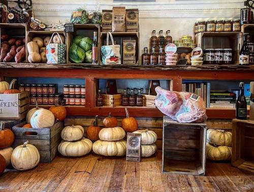 A rustic market display with wooden crates filled with pumpkins, vegetables, jars, and various local products on shelves.