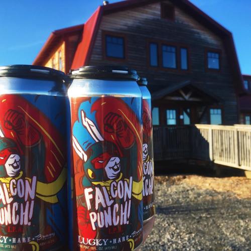 Four cans of "Falcon Punch!" beer in front of a rustic building with a wooden deck and blue sky.