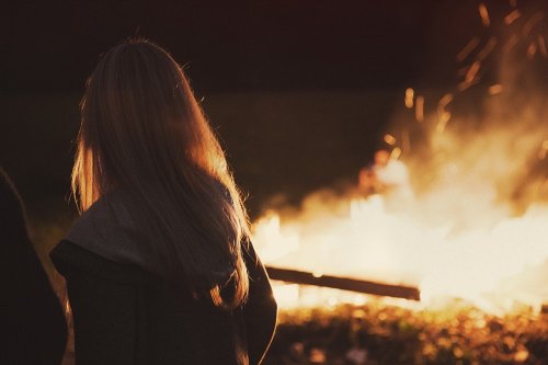 A person with long hair stands in front of a large fire, with sparks flying in the dark background.