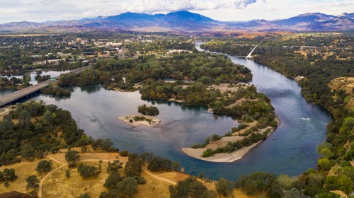 Aerial view of a winding river surrounded by greenery, with mountains in the background and a small island in the water.
