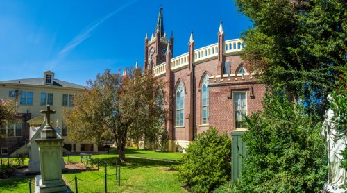 A historic brick church with a clock tower, surrounded by greenery and a cemetery, under a clear blue sky.