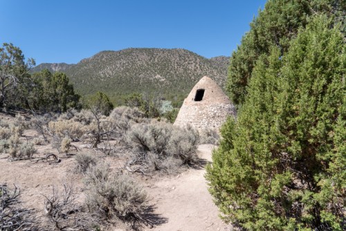 A stone beehive-shaped structure surrounded by shrubs and mountains under a clear blue sky.