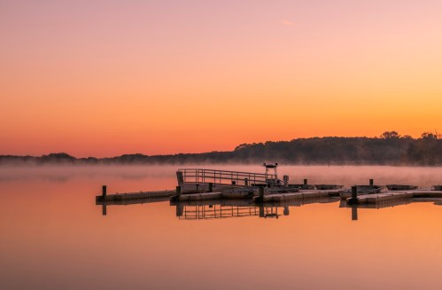 A serene lake at sunrise, with a misty surface and a dock reflecting the warm orange and purple sky.