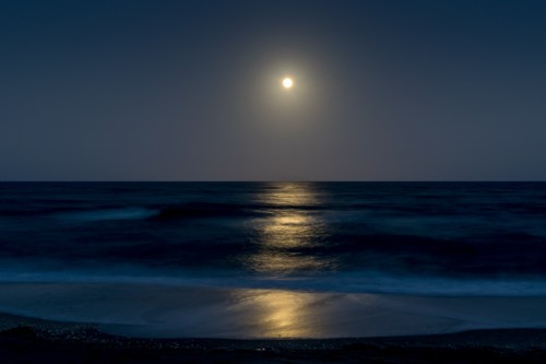 A serene night scene of a full moon reflecting on calm ocean waters, with gentle waves lapping at the shore.