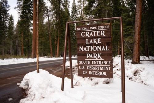 Sign for Crater Lake National Park's south entrance, surrounded by snow and tall evergreen trees.