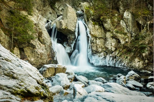 A serene waterfall cascades over rocks, surrounded by icy terrain and lush greenery in a tranquil forest setting.