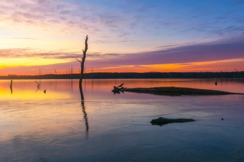 A serene lake at sunset, with colorful skies, silhouetted trees, and reflections on the water's surface.