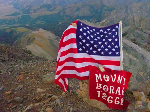 An American flag and a red banner reading "MOUNT BORAH 12662" on a rocky mountain summit with a scenic view.
