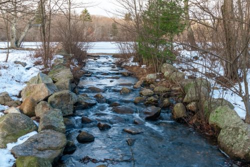 A serene stream flows over rocks, surrounded by snow-covered ground and bare trees in a tranquil winter landscape.