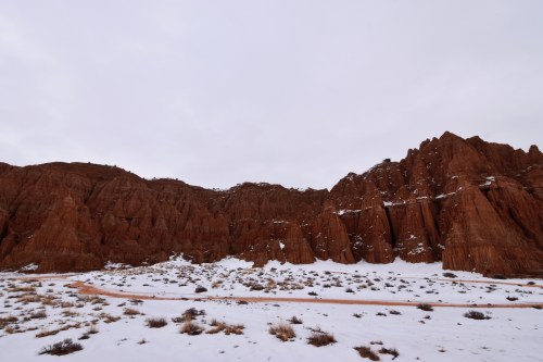 A snowy landscape with red rock formations under a cloudy sky, featuring a winding road in the foreground.