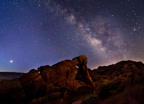 A starry night sky with the Milky Way above rugged rock formations in a desert landscape.