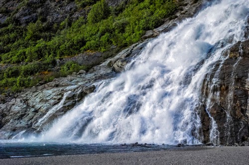A cascading waterfall flows over rocky terrain, surrounded by lush greenery and a clear blue sky.
