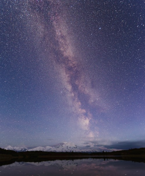 A stunning night sky filled with stars and the Milky Way, reflecting on a calm lake with snow-capped mountains in the background.