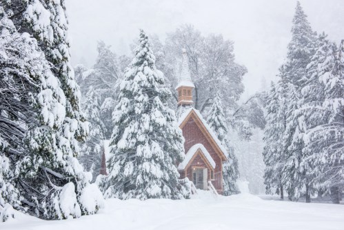 A quaint brick church surrounded by tall, snow-covered trees in a winter landscape. Snowflakes gently fall.