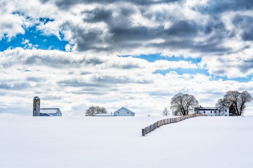 A snowy landscape with a wooden fence, farmhouses, and a silo under a cloudy blue sky.