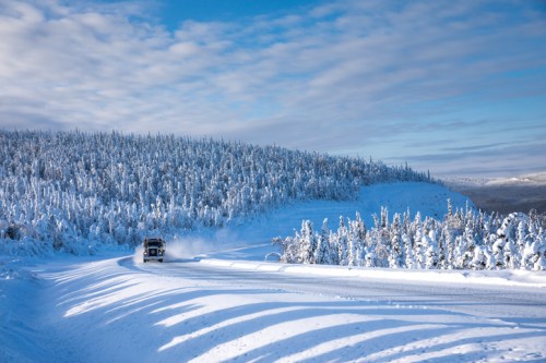 A vehicle drives along a snowy road surrounded by snow-covered trees under a blue sky with scattered clouds.