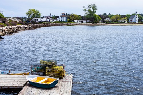 A calm waterfront scene with a yellow boat, lobster traps, and a quaint village in the background under a cloudy sky.