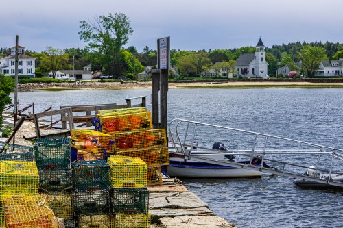 Colorful lobster traps stacked on a dock by a calm bay, with a church and houses visible in the background.