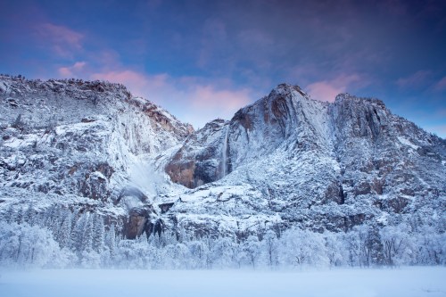 Snow-covered mountains under a colorful sky, with trees blanketed in white, creating a serene winter landscape.