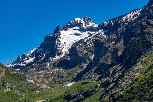 A majestic mountain peak with snow-capped summits, surrounded by rocky cliffs and lush green valleys under a clear blue sky.