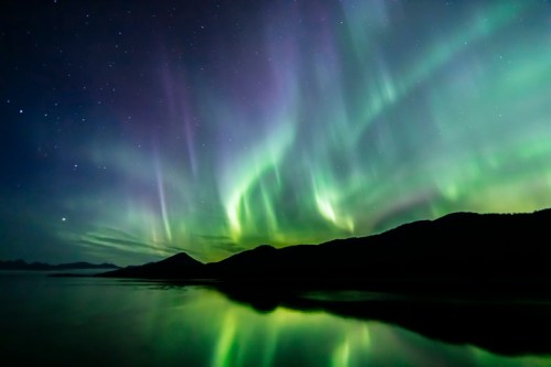 Vibrant auroras dance across the night sky, reflecting on a calm lake with silhouetted mountains in the background.