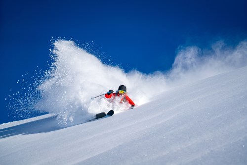 A skier in a red jacket carves through fresh snow, creating a spray against a clear blue sky.