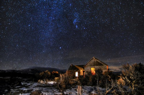 A dilapidated house under a starry night sky, surrounded by sparse vegetation and mountains in the distance.