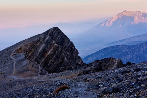 A rugged mountain peak with layered rock formations, overlooking a misty valley and distant mountains at sunrise.