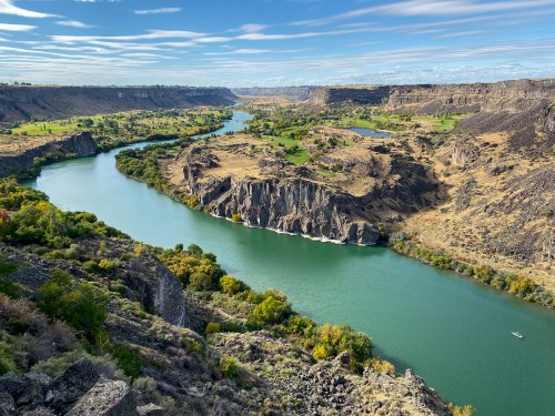 A scenic view of a winding river surrounded by rocky cliffs and lush greenery under a blue sky with scattered clouds.
