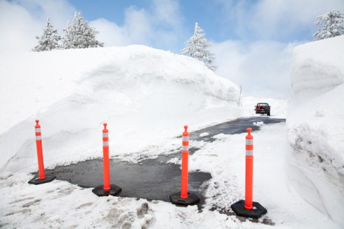 A snow-covered road with tall snowbanks on either side and orange traffic cones marking the path ahead.
