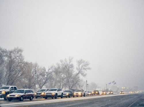 A line of cars on a snowy road, with trees and flags barely visible in the heavy snowfall.