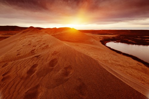 A serene desert landscape at sunset, with sand dunes and a calm body of water reflecting the warm colors of the sky.