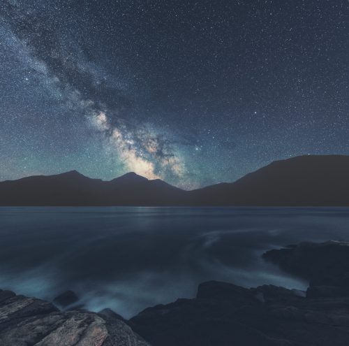 A serene night sky filled with stars and the Milky Way over a calm lake, framed by dark mountains and rocky shore.