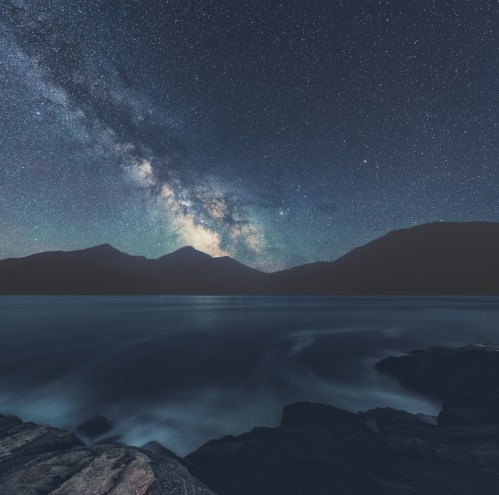 A serene night sky filled with stars and the Milky Way over a calm lake, framed by dark mountains and rocky shoreline.